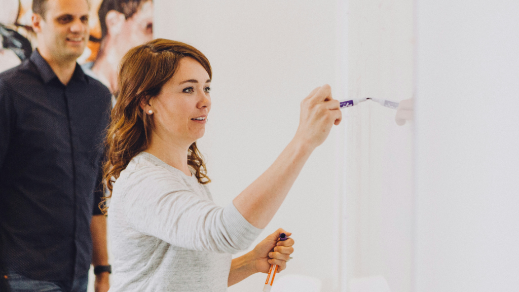 Eine Frau schreibt mit einem lila Marker auf ein Whiteboard, während ein Mann im Hintergrund zuschaut